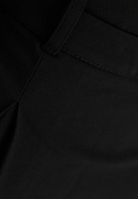 Detailní záběr na černou látku ukazující hladkou strukturu, plochý pas a jemné švy, se slabým leskem od osvětlení.