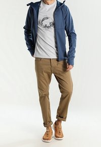 Blå letvægtsjakke med hætte over en grå t-shirt med et elefantgrafik. Tan chinos og tan vævede sneakers fuldender outfittet.