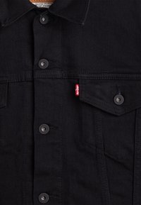 Giubbotto in denim nero con bottoni argentati, tasche con bottone e una etichetta rossa Levi's. Il tessuto ha una texture liscia con cuciture visibili.