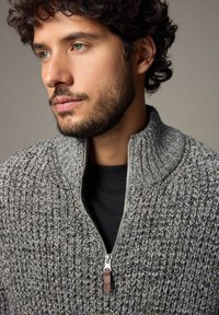 Grå, struktureret zip-sweater med høj krave og en synlig lynlåsdetalje. Bæres over en sort skjorte med mønster.