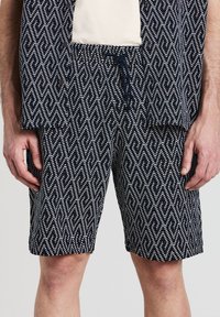 Pantaloni corti blu navy con un motivo geometrico bianco e arancione. Presentano una vita elasticizzata con cordino e tasche laterali. Materiale in cotone.