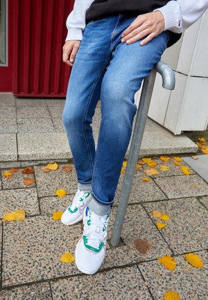 Person, die blaue Jeans und weiße Sneakers trägt, sitzt auf einem Metallrohr auf einem Bürgersteig mit verstreuten gelben Herbstblättern.
