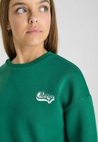 Groene sweatshirt met ronde hals, standaard pasvorm, zachte stof en witte "Cherry" logo op de voorkant. Glad textuur, casual ontwerp.