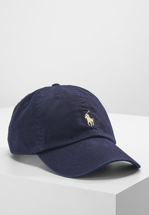 Casquette de baseball en coton bleu marine avec visière courbée et petit logo brodé d'un joueur de polo jaune sur le devant, posée sur une surface blanche.