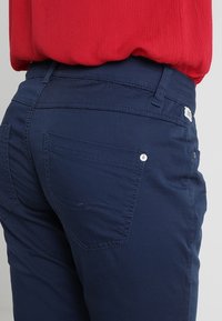 Pantalon bleu marine en mélange de coton, avec une poche arrière munie d'un petit bouton en métal, coupe droite et détails de couture discrets.