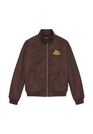 Homme barbu portant une veste marron zippée, un t-shirt blanc, un jean bleu ample et des baskets blanches, debout devant un fond gris.