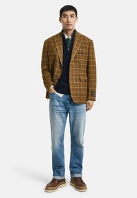 Braun karierter Blazer, marineblauer Pullover, hellblaue Jeans. Braune Schuhe mit beigen Sohlen. Keine weiteren Details oder Muster sichtbar.