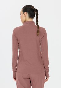 Vrouw met gevlochten haar, gekleed in een mauve langmouw sporttop en bijpassende broek, met de rug naar de camera gericht tegen een eenvoudige witte achtergrond.