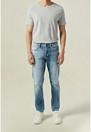 Mann trägt ein hellgraues T-Shirt mit kurzen Ärmeln, ausgewaschene blaue Jeans und weiße Turnschuhe und steht vor einem schlichten neutralen Hintergrund.