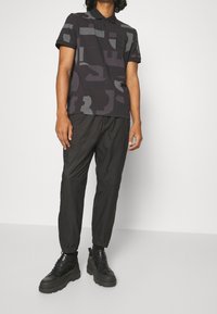 Černé polo tričko s geometrickými vzory, krátkými rukávy a kapsou. V kombinaci s černými zúženými kalhotami a platformovými teniskami.