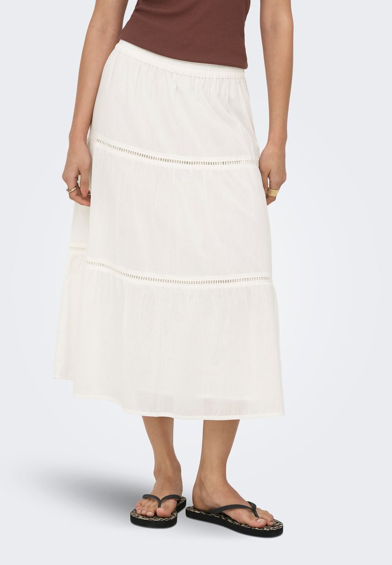 Femme portant une jupe midi blanche à volants, un haut marron sans manches et des sandales tongs noires avec des semelles à motifs.