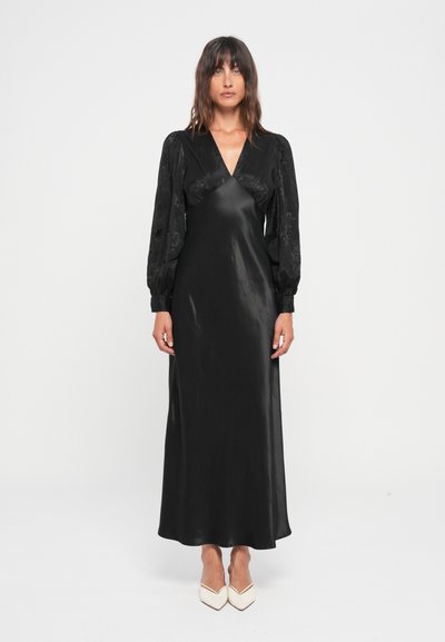 Vestido largo de satén negro con escote en V y mangas largas abullonadas que presentan un patrón floral texturizado. Combinado con tacones blancos de punta.