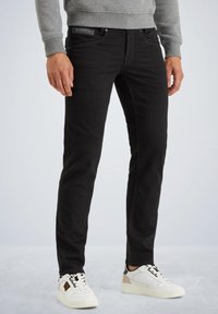 Čierne denimové džínsy so slim fit strihom, s prednými vreckami a štítkom na páse, v kombinácii s bielymi teniskami s čiernymi a béžovými akcentmi.