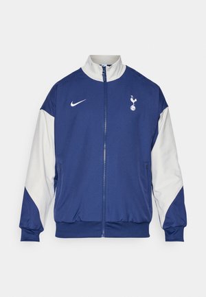 Tumši zilas krāsas jaka ar baltām piedurknēm, pilna zīmogojuma aizdare, augsts apkakles dizains, Nike logo un Tottenham Hotspur emblēma kreisajā krūtī.