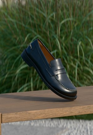 Donkerblauwe leren loafer met beige stiksels, tentoongesteld op een houten oppervlak tegen een achtergrond van hoog groen gras.