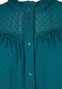 Blouse sarcelle en tissu à pois texturé, col à volants et empiècement, patte de boutonnage sur le devant, et détail en dentelle sur la poitrine.