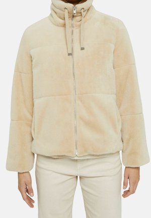 Veste en fausse fourrure beige avec un col haut, une fermeture éclair devant, des cordons de serrage et des manches longues, portée avec un pantalon clair.