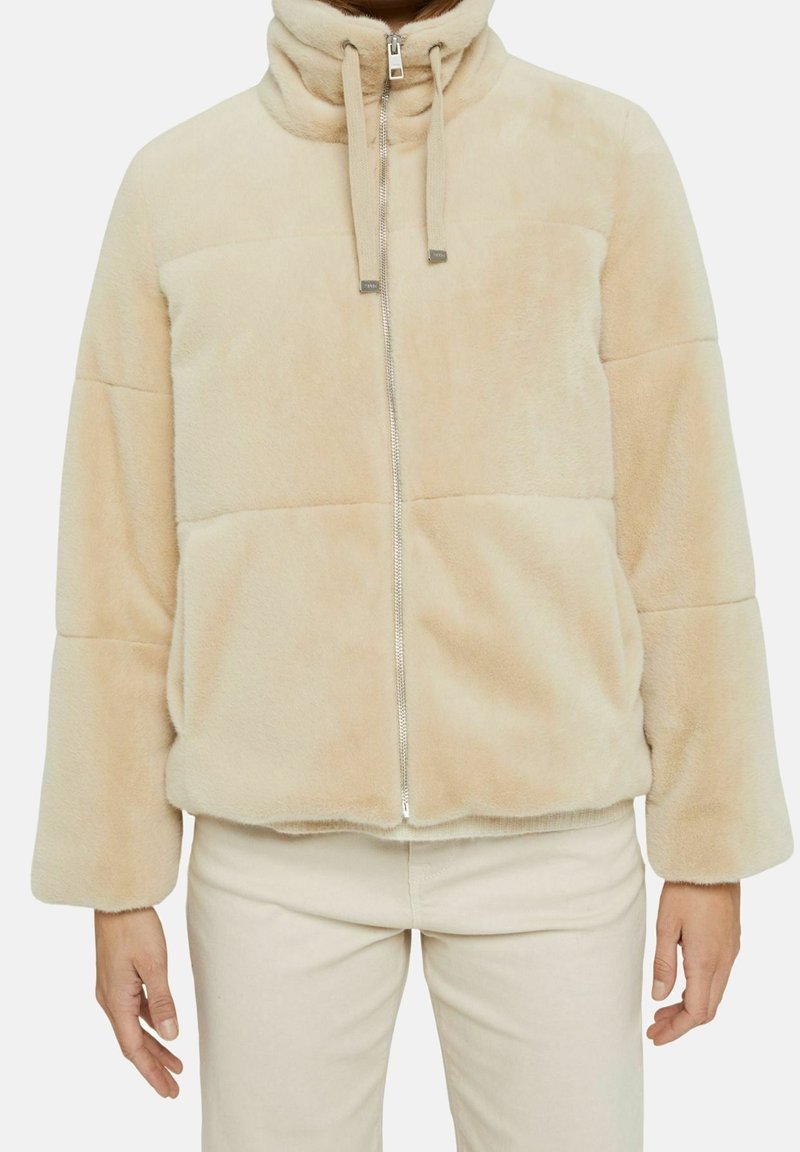 Veste en fausse fourrure beige avec un col haut, une fermeture éclair devant, des cordons de serrage et des manches longues, portée avec un pantalon clair.