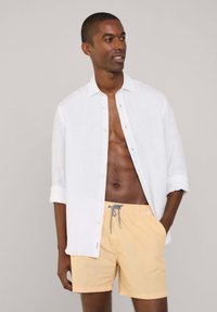 Chemise en lin blanche avec manches retroussées sur un short de bain rayé jaune, doté d'une taille élastique et d'un cordon de serrage.