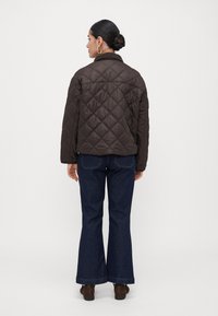Woman with hair in bun wearing gold hoop earrings, dark brown quilted jacket, dark blue flared jeans, and dark brown shoes, facing away.