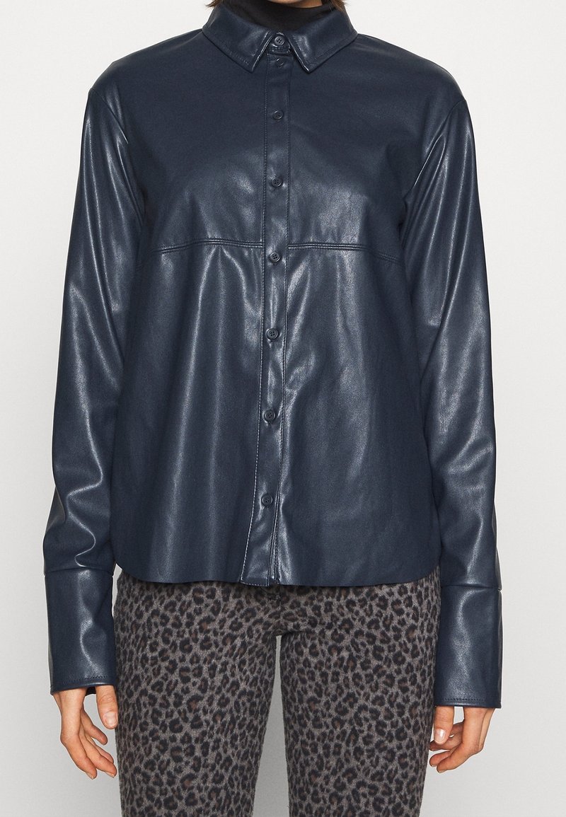 Chemise à boutons en similicuir bleu marine avec col, présentant une texture lisse et des coutures apparentes, portée avec un pantalon à imprimé léopard.