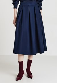 Gonna midi plissettata blu mare realizzata in tessuto morbido con una vita aderente. Indossata con calze a coste bordeaux e tacchi lucidi bordeaux.