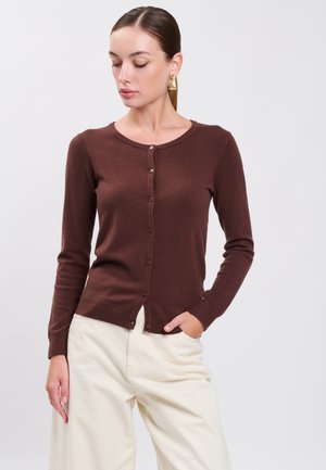 Femme portant un cardigan marron boutonné et un pantalon blanc, debout avec une main dans la poche et regardant vers le bas sur le côté.