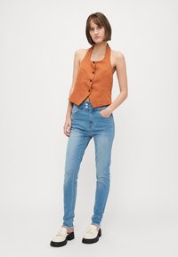Rozsdabarna, nyakpántos mellény gombos elejével, világoskék skinny farmerral párosítva. Fehér, fekete talpú slip-on cipők. Minimalista dizájn.
