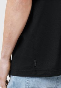 T-shirt nera a maniche corte, con una texture liscia e un'etichetta visibile sulla cucitura laterale. Indossata con jeans azzurri chiari.