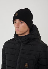 Giacca a vento nera con chiusura a zip, cuciture orizzontali e tasca sul petto. Abbinata a un berretto a coste nero con un'etichetta logata.