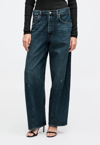 Modell trägt dunkelblaue, weit geschnittene Jeans aus Denim, kombiniert mit schwarzen, schmalen High-Heel-Sandalen und einem langärmligen schwarzen Oberteil.