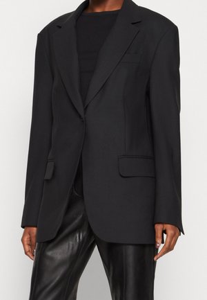 Persoon draagt een zwarte blazer met een enkele knoop en klepzakken, gecombineerd met zwarte leren broek en een zwart topje eronder.