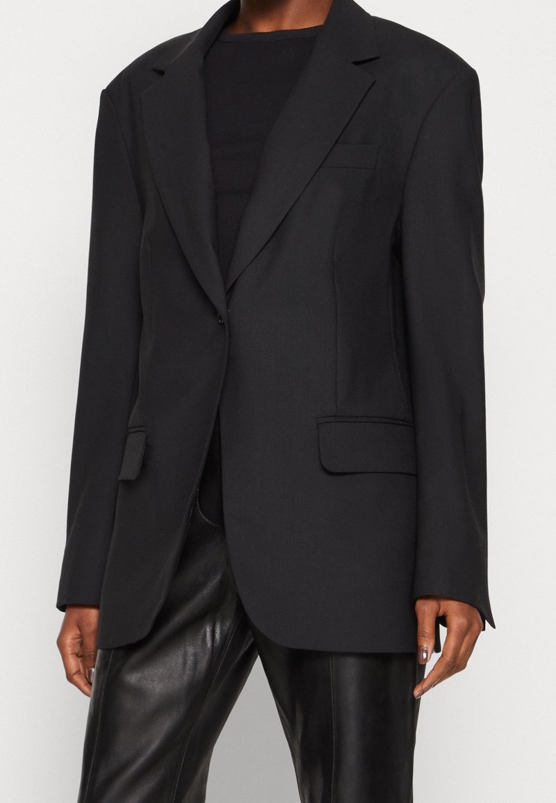 Personne portant un blazer noir à un seul bouton avec poches à rabat, associé à un pantalon en cuir noir et un haut noir en dessous.