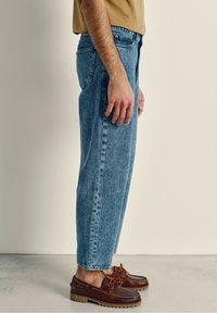 Svetlo modri jeans s ravnim krojem, vidnimi šivi, stranskimi žepi in visokim pasom, v kombinaciji z rjavimi usnjenimi čolnarskimi čevlji.