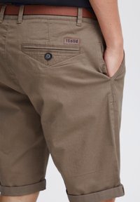 Pantalones cortos de algodón marrón con puños enrollados, bolsillos traseros y una etiqueta de cuero que dice "Solid". Tela texturizada con un brillo sutil.