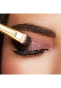 En guld makeupborste applicerar glittrande plommonögonskugga på ett stängt ögonlock, med mörka ögonfransar och väldefinierad ögonbryn. Slät hudstruktur synlig.