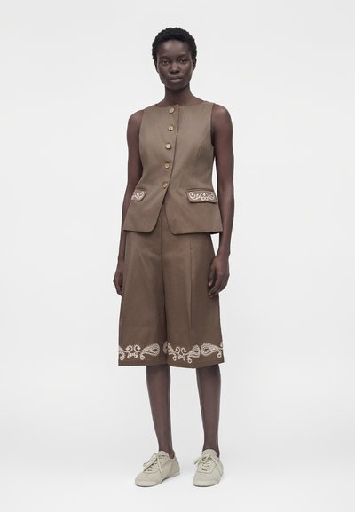 Γυναίκα που φορά αμάνικο καφέ γιλέκο με κουμπιά και αντίστοιχα σορτς μέχρι το γόνατο με λευκή κεντητική, συνδυασμένα με ανοιχτό μπεζ αθλητικά παπούτσια.