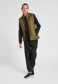 Olivgrön vadderad väst med hög krage, dragkedja fram, bärs över en svart långärmad topp och svarta byxor, kompletterad med svarta texturerade sneakers.