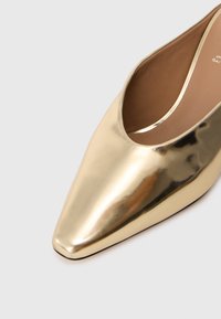 Złote metaliczne buty na obcasie, zakończone szpicem, z gładką powierzchnią i zakrzywionym wycięciem po bokach, ukazujące gładką teksturę oraz dopasowane wnętrze.