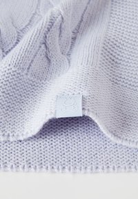 Svetle fialová pletená deka s texturovaným vzorem, která má měkký, huňatý povrch a malou modrou textilní značku na okraji.