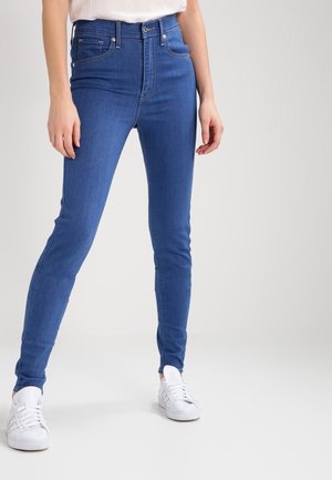 Persoon die nauwsluitende blauwe spijkerbroek en witte sneakers draagt, staat tegen een eenvoudige lichte achtergrond.
