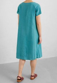 Türkises Leinenkleid, kurze Ärmel, lockere Passform, runder Halsausschnitt, knielange Länge, mit einer geraden Naht auf der Rückseite, kombiniert mit braunen Sandalen.