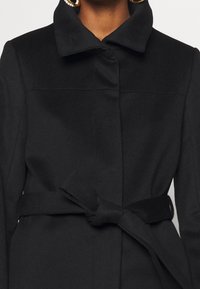 Černý kabát s strukturovaným límcem, předním zapínáním na knoflíky a textilním opaskem uvázaným v pase. Měkká textura s minimalistickými designovými detaily.