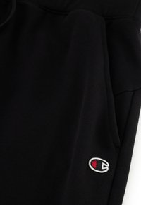 Zwarte trainingsbroek van zacht materiaal, met een elastische tailleband en zijzakken. Geborduurd logo in het wit en rood op het linkerbeen.