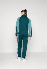 Tamsiai žalia sportinė apranga su šviesiai mėlynomis detalėmis, pasižyminti aukšta apykakle ir juostelėmis, derinama su baltais sportiniais batais.