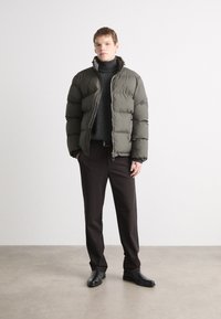 Jachetă puffer de culoare verde măslinie cu guler înalt, combinată cu un pulover negru cu guler înalt și pantaloni maro, purtată cu botine negre elegante.