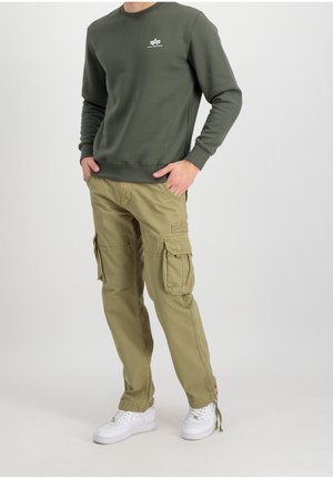 Mand iført olivengrøn Alpha Industries sweatshirt, khakifarvede cargobukser med flere lommer og hvide sneakers, stående med hænderne i lommerne.