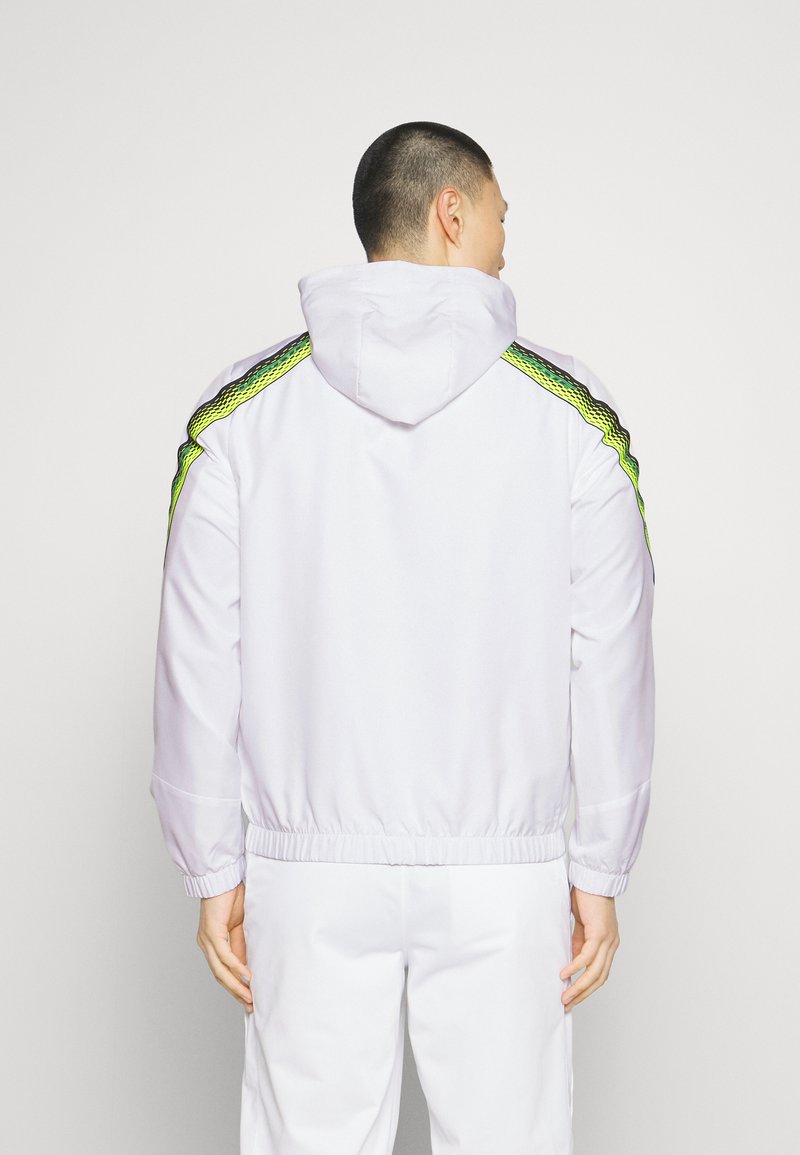 Homme aux cheveux courts portant une veste à capuche blanche avec des rayures vert fluo et noires sur les manches, se tenant dos à la caméra.