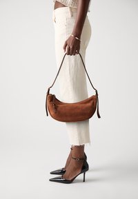 Borsa in suede marrone con forma curvata, manico corto in pelle e dettagli di cucitura sottili. Modella che indossa pantaloni crema e tacchi neri.