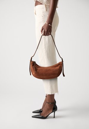 Borsa in suede marrone con forma curvata, manico corto in pelle e dettagli di cucitura sottili. Modella che indossa pantaloni crema e tacchi neri.